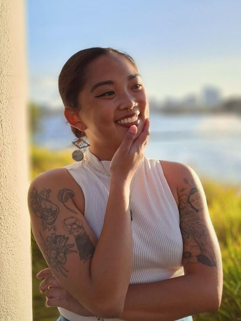 Annelle smiling, wearing a white sleeveless turtleneck and gold and abalone earrings. They have their hand on their chin and have tattoos on their upper arms.