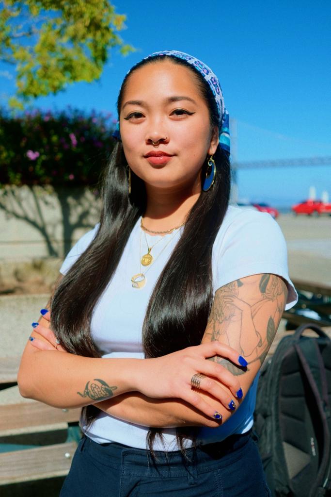 Annelle looking at the camera, arms crossed, wearing a white tee shirt and blue bandana. Behind them is the Bay Bridge.