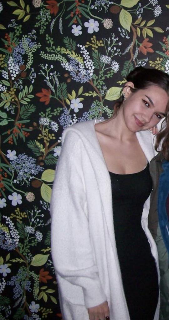 Colby in a black dress, white long cardigan, leaning on a friend who is cropped out with a floral wallpaper backdrop. 