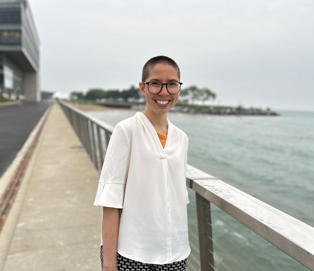 Maddie standing and smiling in front of a lake. They have a buzz cut and glasses and are wearing a white shirt.
